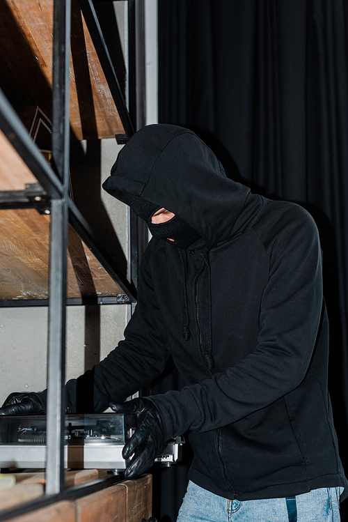 Selective focus of robber in mask and leather gloves taking record player from cupboard