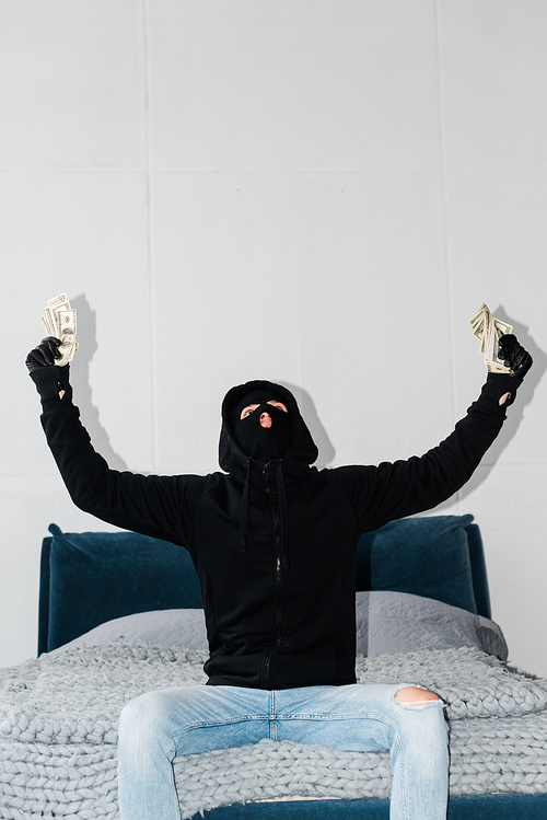 Robber in balaclava and leather gloves holding dollars on bed