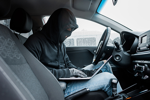 Side view of robber in balaclava and leather gloves using laptop while sitting in car