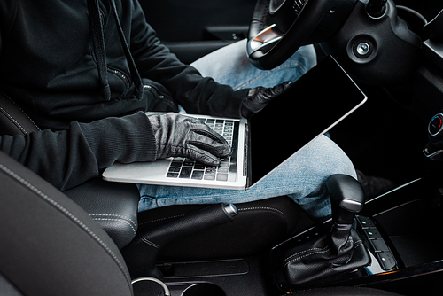Cropped view of thief in leather gloves using laptop with blank screen in auto