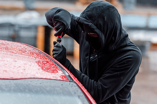 Selective focus of robber in mask and leather gloves opening door of auto with screwdriver outdoors
