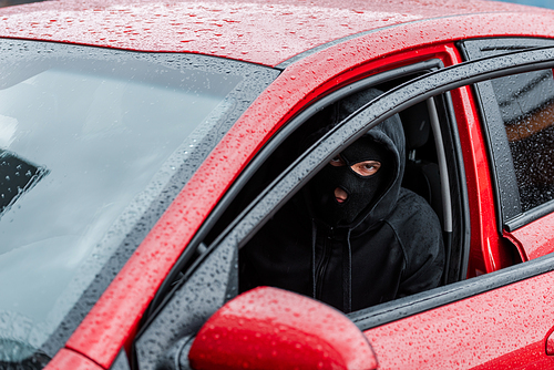 Selective focus of robber in balaclava  while sitting in car with open door