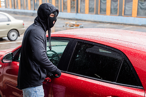 Robber in balaclava and leather gloves looking away while opening car with screwdriver on urban street