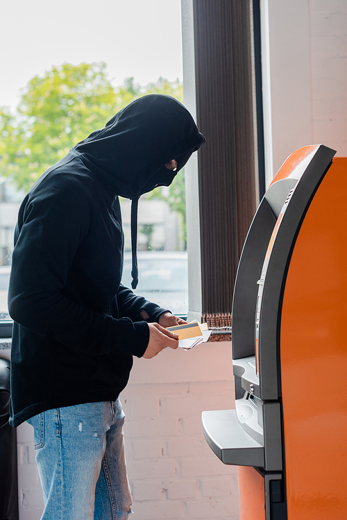 Side view of robber in balaclava holding credit cards near atm