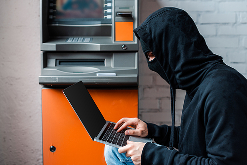Side view of hacker in mask using laptop with blank screen near atm