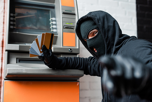 Selective focus of burglar in balaclava pointing with finger at camera while holding credit cards near atm