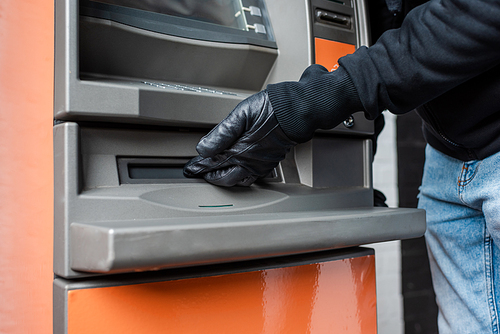 Cropped view of burglar in leather glove holding hand near cash dispenser of automated teller machine