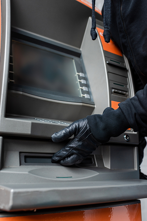 Cropped view of thief in leather glove holding hand near cash dispenser of atm