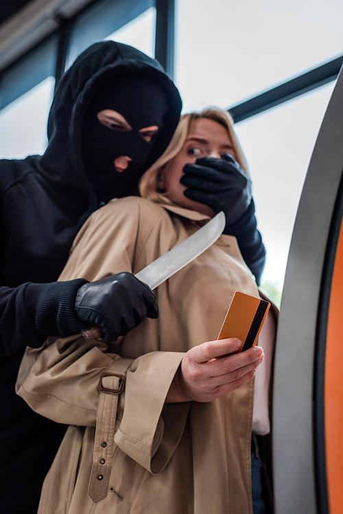 Low angle view of robber holding knife and covering mouth to scared woman holding credit card near atm
