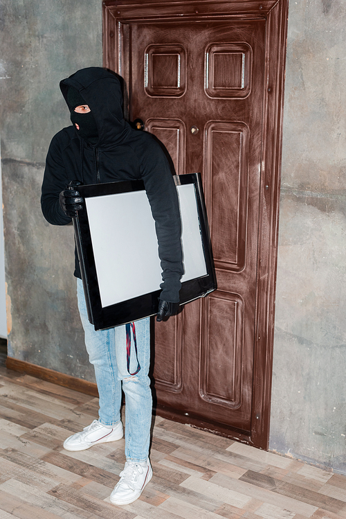 Thief in balaclava and leather gloves holding tv near house door