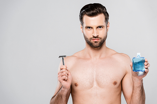 bearded and muscular man holding after shave lotion and razor isolated on grey