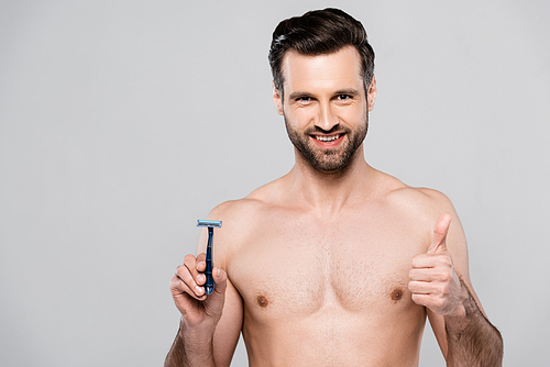 handsome man holding razor while smiling and showing thumb up isolated on grey