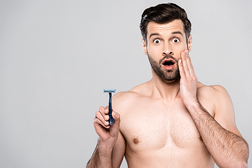 shocked and bearded man holding razor while touching face isolated on grey