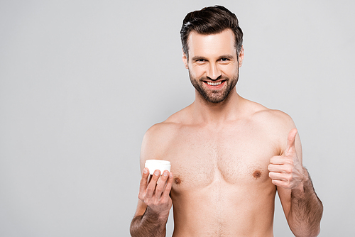 happy bearded man holding container with cosmetic cream and showing thumb up isolated on grey