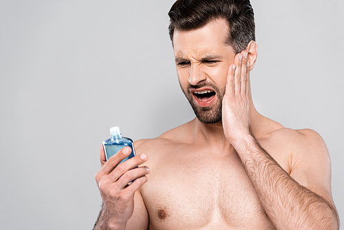 muscular man touching face and looking at after shave lotion isolated on grey
