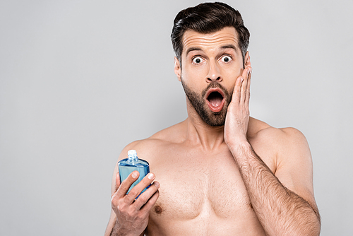 shocked man holding bottle with after shave lotion and touching face isolated on grey