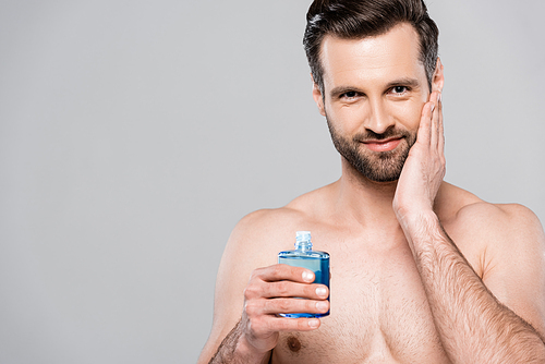 happy man holding bottle with blue after shave lotion and touching face isolated on grey