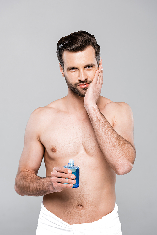 muscular man holding bottle with blue after shave lotion and touching face isolated on grey
