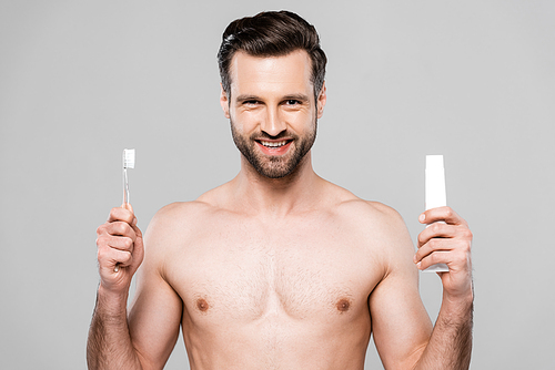 happy and muscular man holding toothpaste and toothbrush isolated on grey