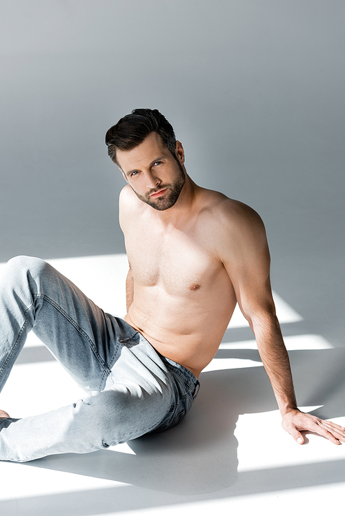 sunlight on muscular and shirtless man in denim jeans sitting on grey