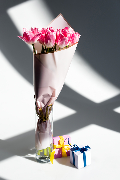 sunlight on pink tulips in vase near gift boxes on white, mothers day concept