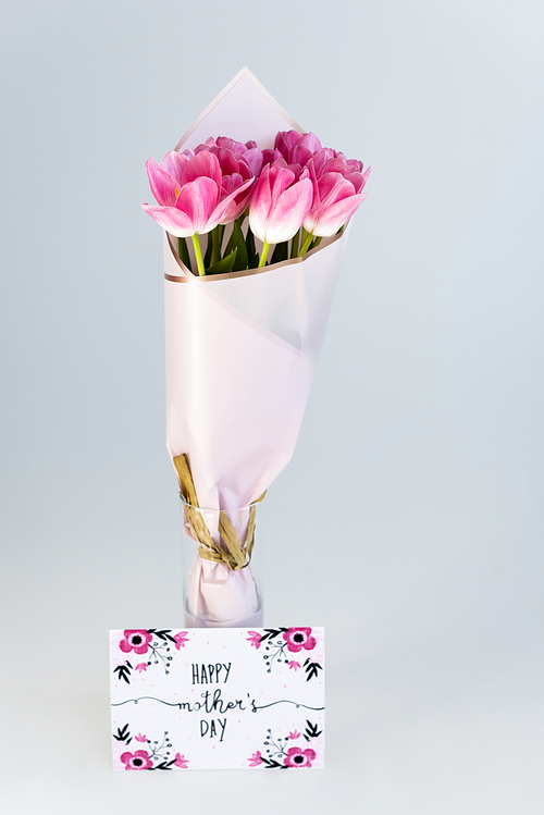 pink tulips in vase near greeting card with happy mothers day lettering isolated on grey