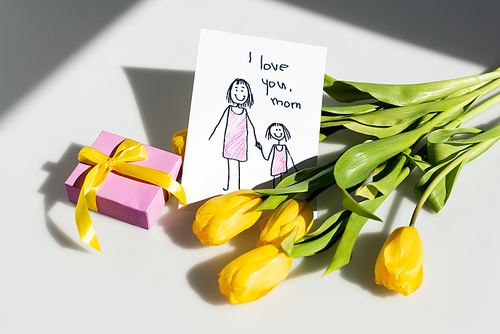 sunlight on yellow tulips near gift box and greeting card with i love you mom lettering on white, mothers day concept