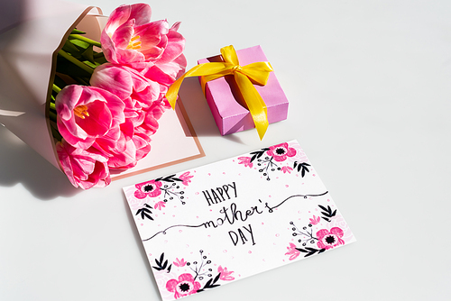 sunlight on pink tulips near gift box and greeting card with happy mothers day lettering on white