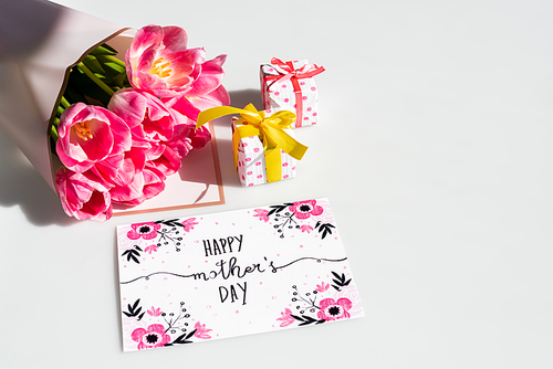 bouquet of tulips near small gift boxes and greeting card with happy mothers day lettering on white