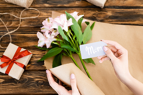 top view of woman holding tag with mom lettering near pink flowers and gift box on wooden surface, mothers day concept