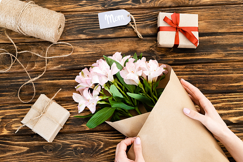cropped view of woman wrapping pink flowers in paper near gift boxes on wooden surface, mothers day concept