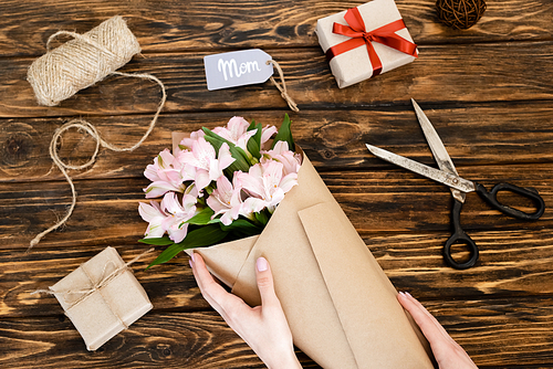 cropped view of woman touching wrapped in paper pink flowers near gift boxes and tag with mom lettering on wooden surface, mothers day concept