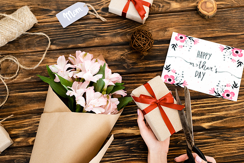 cropped view of woman cutting ribbon on present near pink flowers, greeting card with happy mothers day and tag with mom lettering on wooden surface