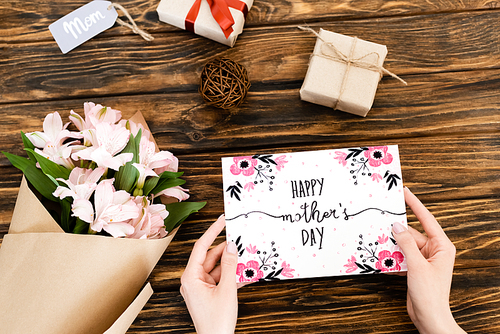 cropped view of woman holding greeting card with happy mothers day lettering near flowers and presents on wooden surface