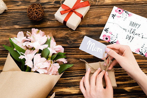 cropped view of woman touching tag with mom lettering on gift box near pink flowers and greeting card with happy mothers day on wooden surface