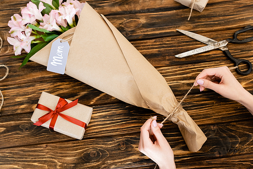 cropped view of woman touching jute twine rope on pink flowers wrapped in paper near gift box and scissors, mothers day concept