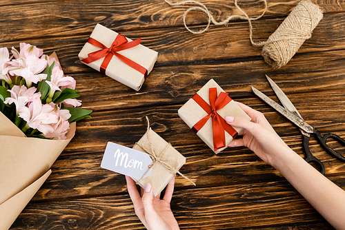 cropped view of woman holding gift boxes near pink flowers wrapped in paper and jute twine rope on wooden surface, mothers day concept