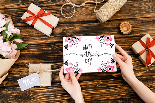 cropped view of woman holding greeting card with happy mothers day lettering near presents and flowers on wooden surface