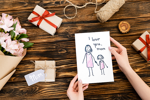 cropped view of woman holding greeting card with i love you mom lettering near presents and flowers on wooden surface