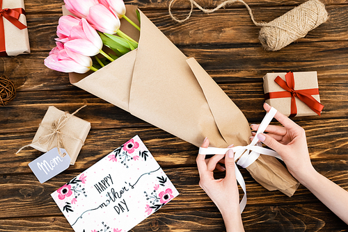 cropped view of woman touching ribbon on wrapped in paper pink tulips near greeting card with happy mothers day lettering and gift boxes on wooden surface