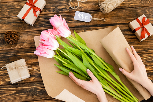 cropped view of woman wrapping pink tulips in paper near gift boxes on wooden surface, mothers day concept