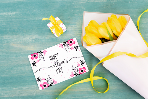 top view of small gift box near greeting card with happy mothers day lettering and yellow tulips on blue wooden surface