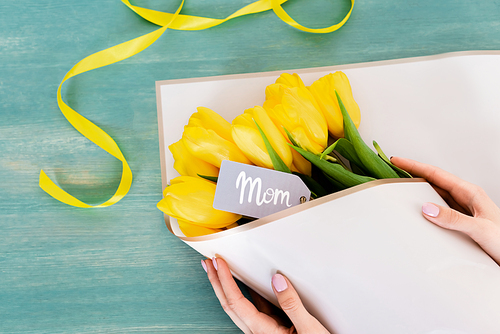cropped view of woman touching wrapped in paper yellow tulips with mom tag lettering on blue surface, mothers day concept