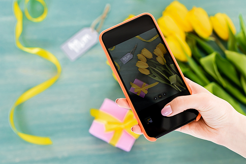 selective focus of woman taking photo of yellow tulips, gift box and mom tag lettering  on textured surface, mothers day concept