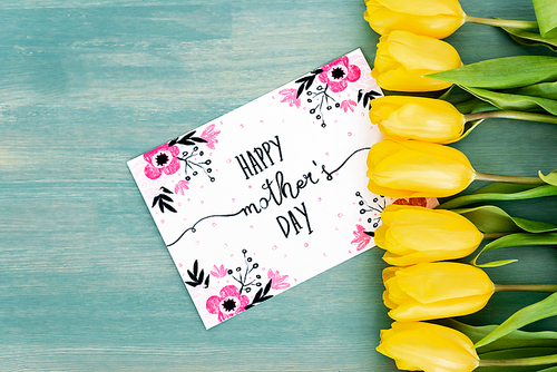 top view of greeting card with happy mothers day lettering near yellow tulips on blue textured surface