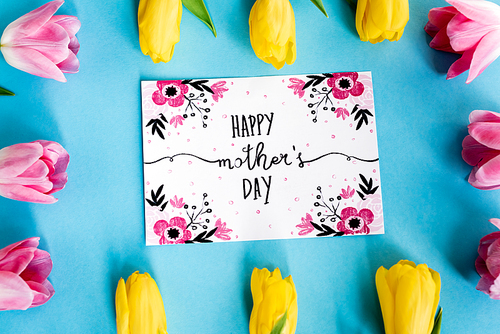 top view of blooming yellow and pink tulips near greeting card with happy mothers day lettering on blue