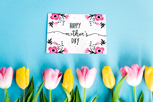 top view of yellow and pink tulips near greeting card with happy mothers day lettering on blue