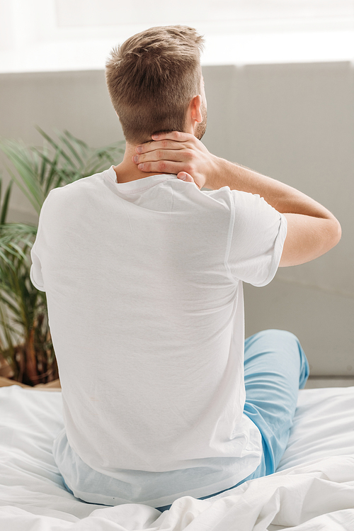 back view of man sitting on white bedding and suffering from neck pain