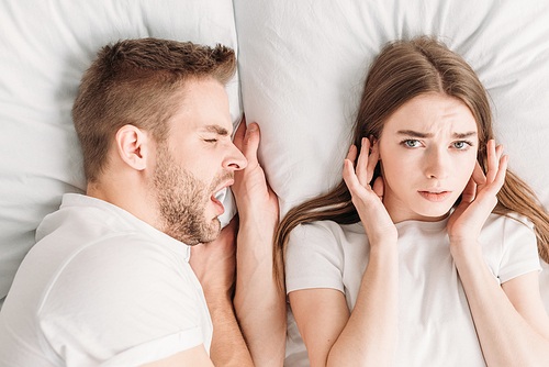 top view of dissatisfied woman  ears with hands and  while lying in bed near snoring husband