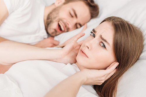 dissatisfied woman  ears with hands and  while lying in bed near snoring husband